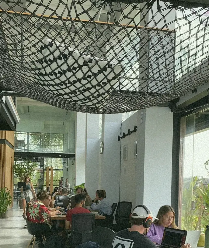 A bustling coworking space at at06 in Canggu, with digital nomads working at a communal table under a large hammock net, fostering a sense of community in Bali.