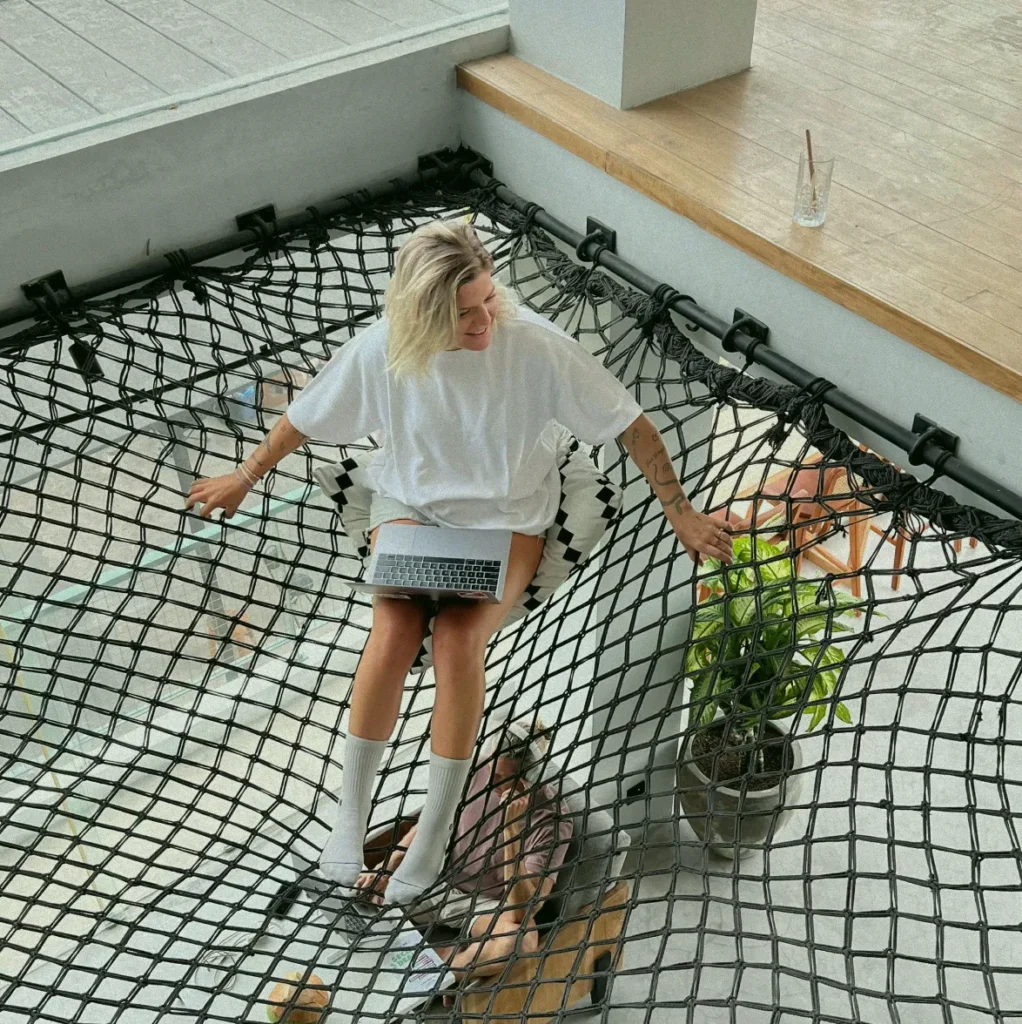 A smiling digital nomad working on her laptop while sitting on a large hammock net at the at 06 coworking space in Canggu, a unique feature of this Bali wellness hub.
