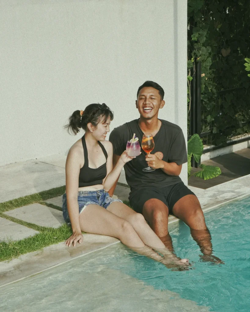 A laughing couple enjoying refreshing cocktails by the poolside at 06 in Canggu, Bali, showcasing a joyful and relaxed wellness experience.
