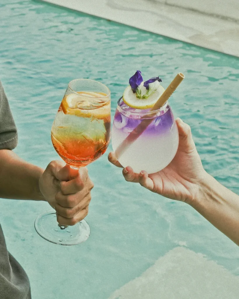 Two hands holding vibrant, refreshing cocktails by the poolside at 06 in Canggu, Bali, perfect for a relaxing wellness experience.