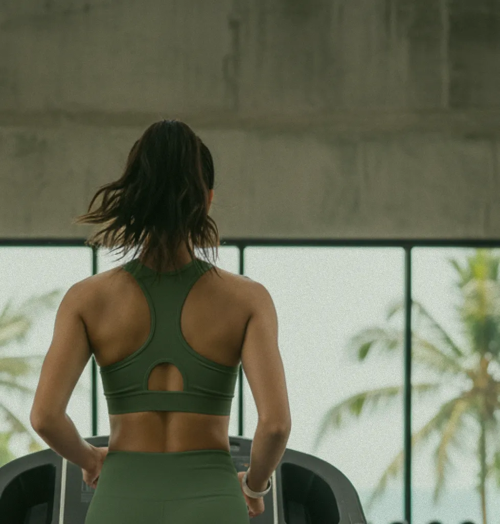 A woman running on a treadmill at at06 gym in Canggu, Bali, with palm trees visible through the window, promoting a healthy lifestyle.