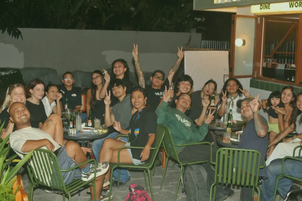 A large group of diverse young adults socializing and making peace signs at an outdoor night event at at06 in Canggu, Bali.