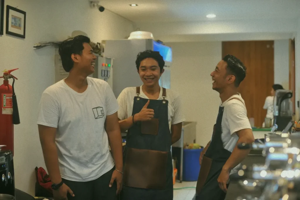 Three friendly baristas laughing together in the kitchen of a Canggu cafe, creating a welcoming atmosphere.