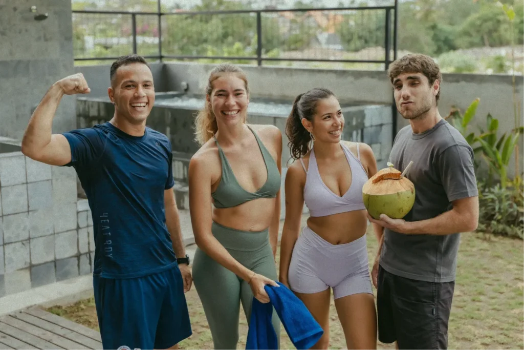 A group of friends in workout clothes posing and smiling after a session at a Canggu gym, one holding a fresh coconut.
