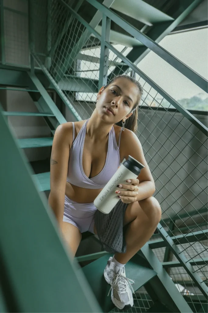 A woman in workout clothes crouching on the stairs of a Canggu gym, holding a water bottle.