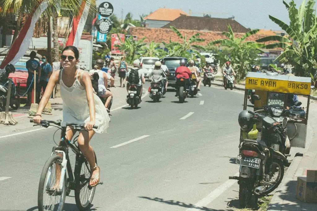 A woman cycling through the bustling streets of Canggu, Bali, with motorbikes and a food stall in the background, capturing the vibrant local life.