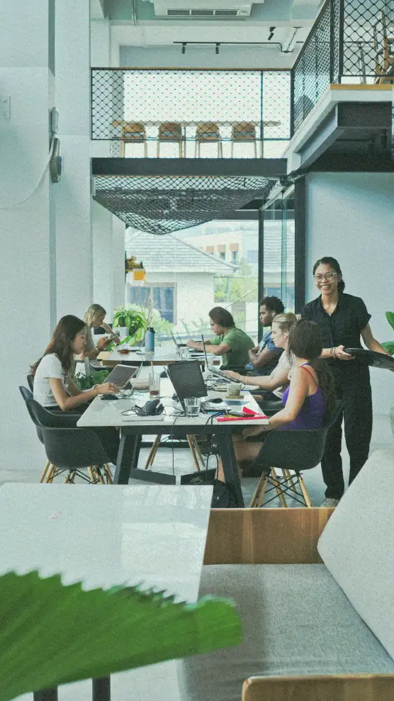 Digital nomads working at a communal table in a bright, modern coworking space in Canggu, with a smiling staff member providing service.