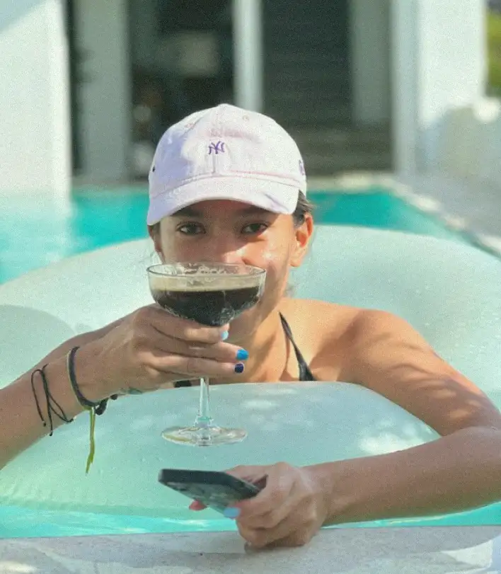 A woman in a pool, wearing a light pink NY baseball cap, sipping a dark cocktail, and holding a smartphone, embodying relaxation and leisure at a Bali wellness hub.