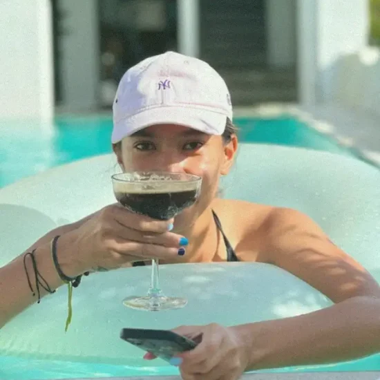 A woman in a pool, wearing a light pink NY baseball cap, sipping a dark cocktail, and holding a smartphone, embodying relaxation and leisure at a Bali wellness hub.